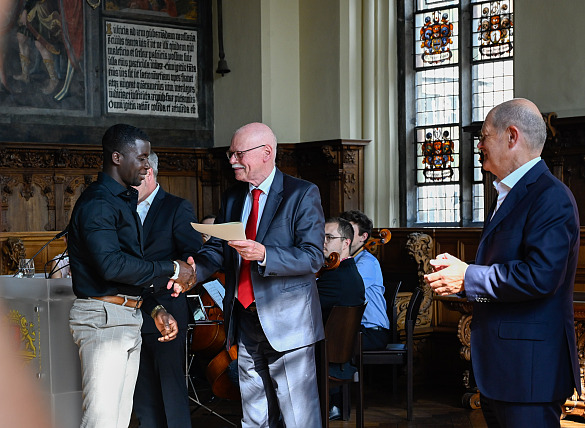 Innensenator Ulrich Mäurer überreicht einem neu eingebürgerten Bremer seine Urkunde; auch Bundeskanzler Scholz (re.) gratuliert. Foto: Senatspressestelle