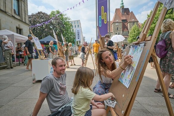 Eine Familie malt gemeinsam auf dem belebten Platz vor der Kunsthalle Bremen im Rahmen des Projekts Kunst vor der Halle, umgeben von vielen Staffeleien und kreativen Köpfen. 